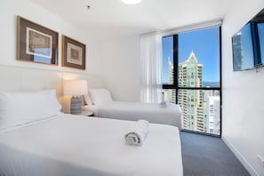 A hotel room with two beds, a nightstand, a lamp, a flat-screen TV, and a large window with a city view. - Rhapsody Stunning Ocean Views Apartment (Surfers Paradise)