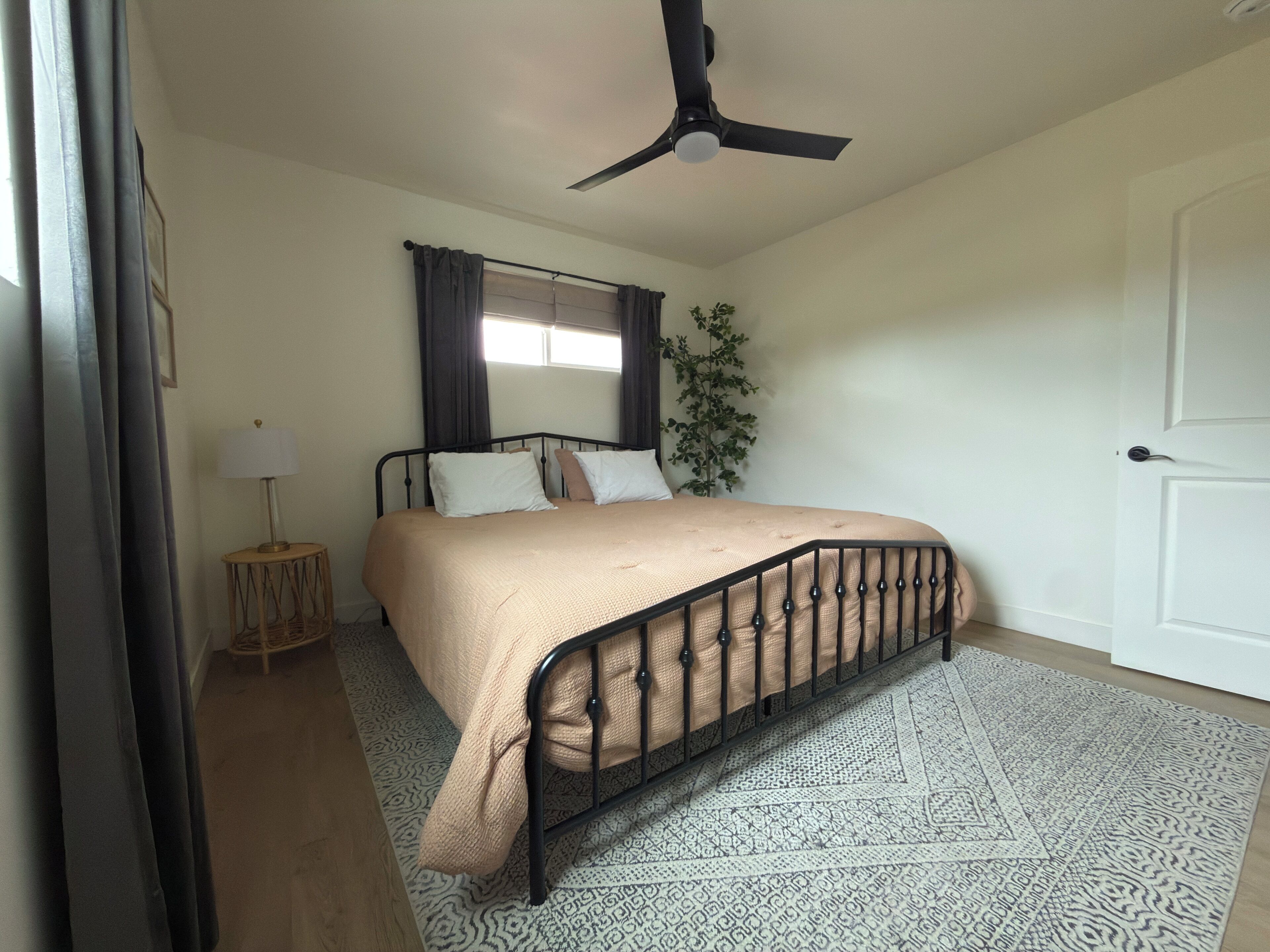 Quarto com cama, ventilador de teto, janela com cortinas, luminária de cabeceira e tapete estampado.
