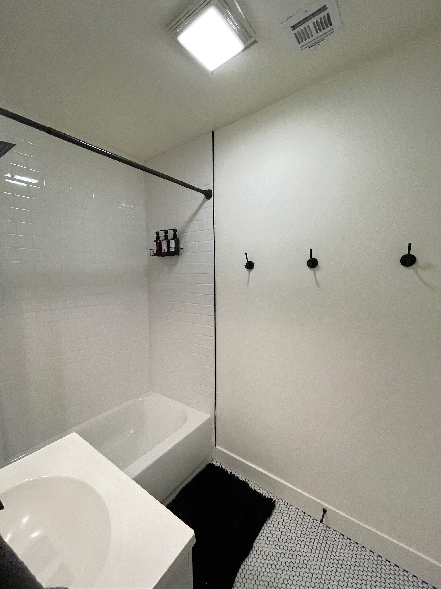 Uma casa de banho moderna com uma banheira branca, uma pia branca e zona de duche. Há três ganchos pretos para toalhas na parede.