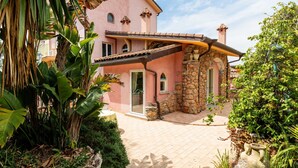 Uma villa rosa com um arco de pedra, um pátio ladrilhado e vegetação exuberante.