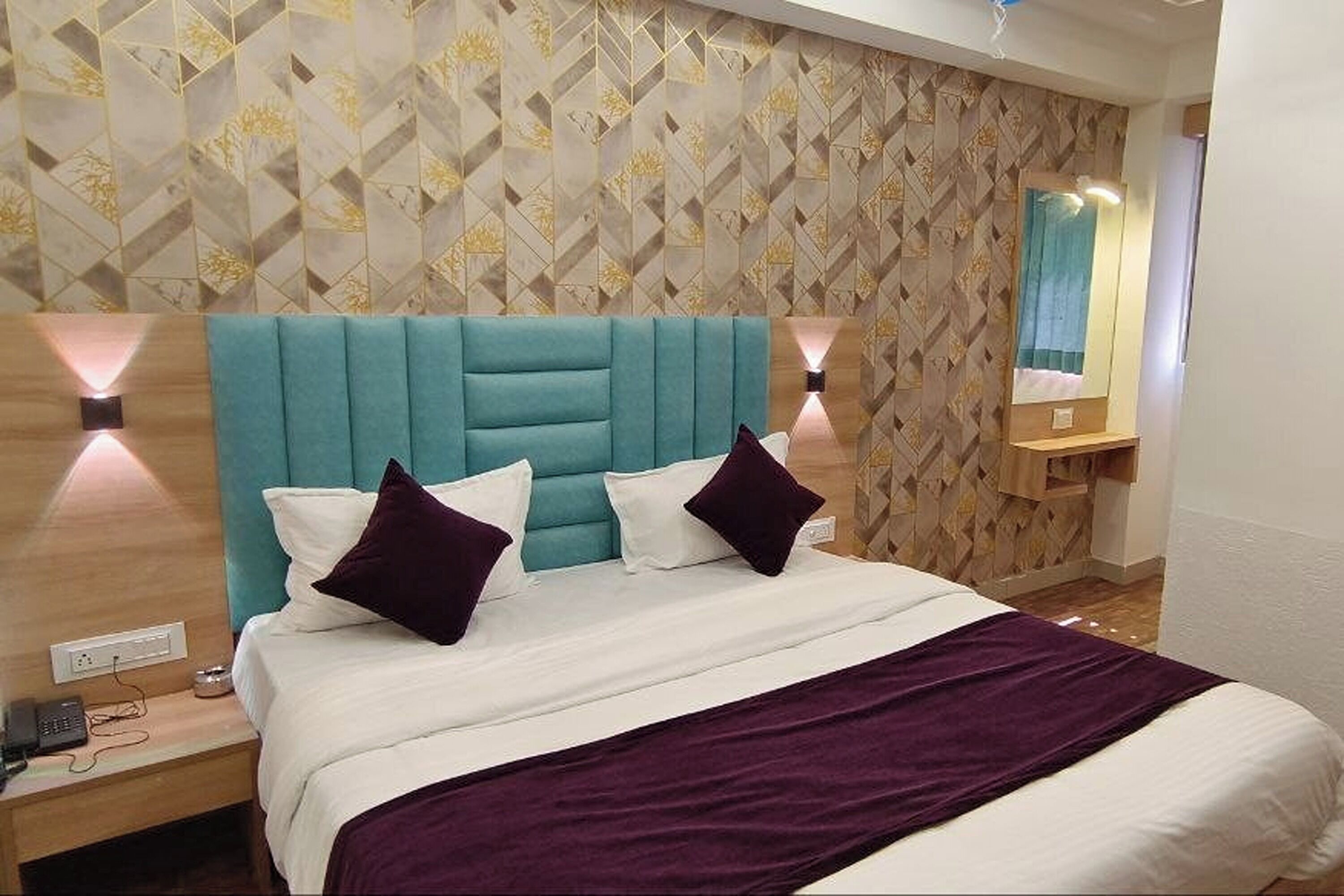 A hotel room with a teal upholstered headboard, white bedding, and purple throw pillows. There are two wall-mounted lamps on either side of the bed, and a wooden nightstand with a phone and a lamp.