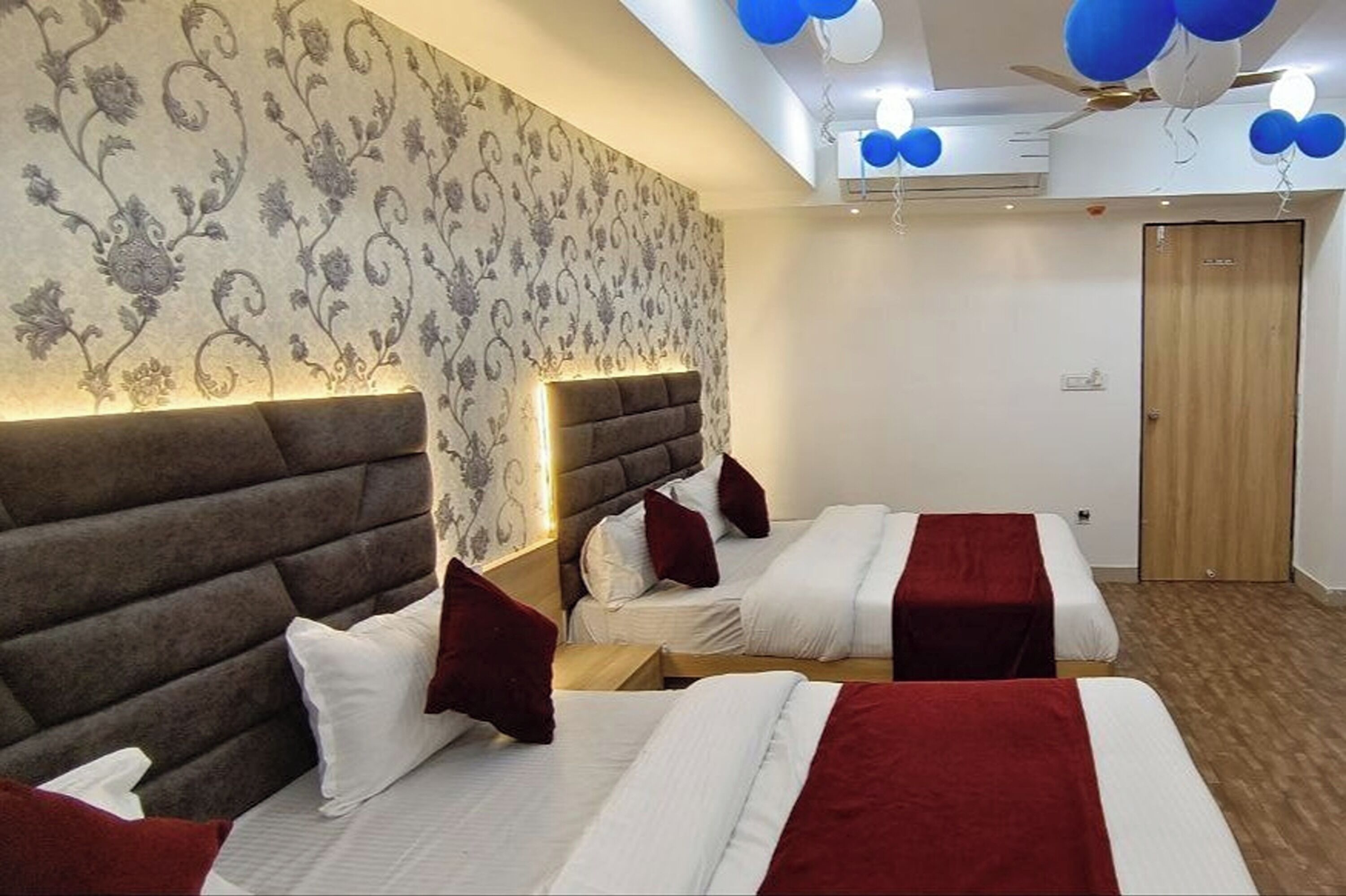 Ein Hotelzimmer mit drei Betten, einem gemusterten Wandteppich, einem Deckenventilator und einer Holztür.