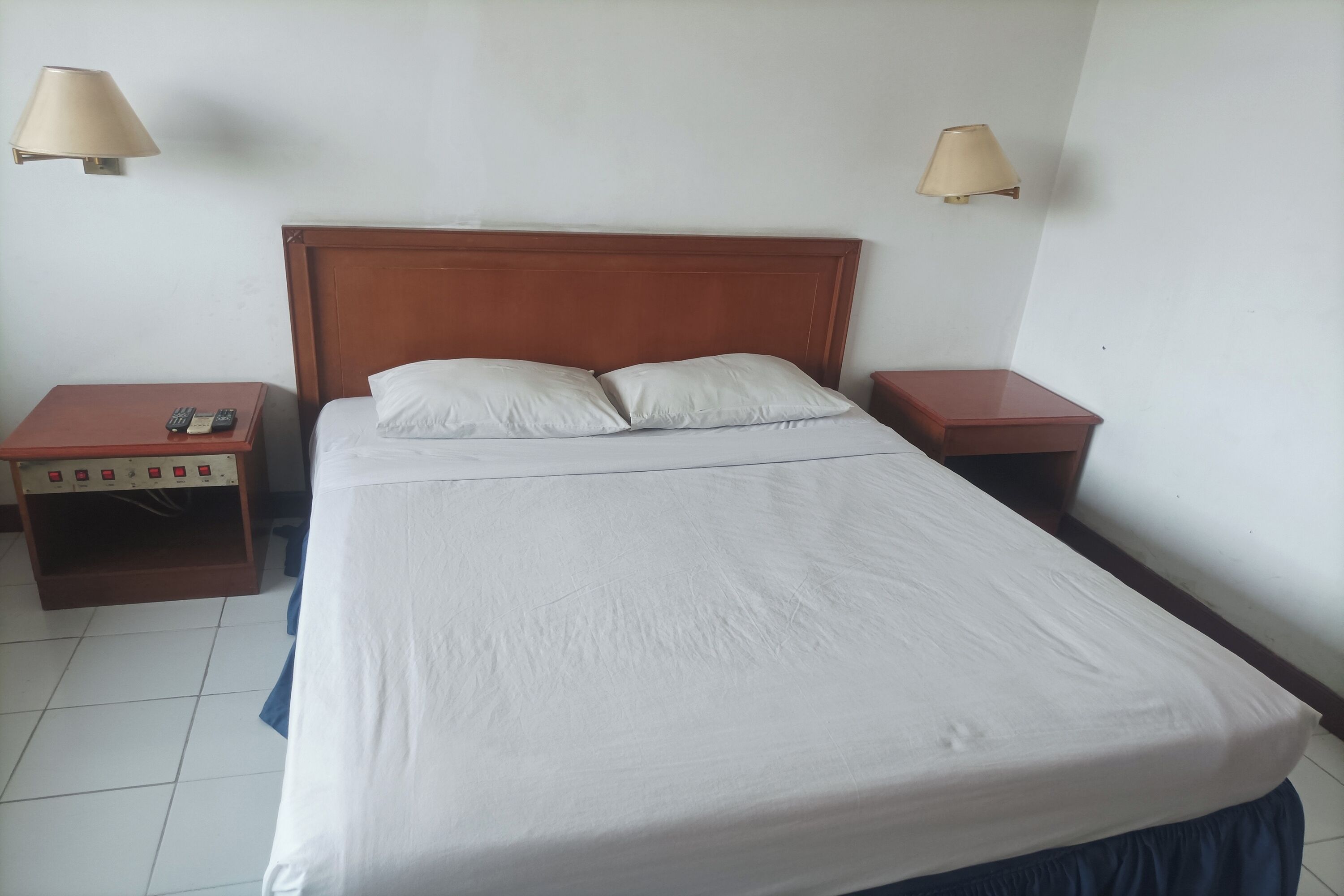 A hotel room with a wooden headboard, a white bedspread, two bedside tables, and two wall-mounted lamps.