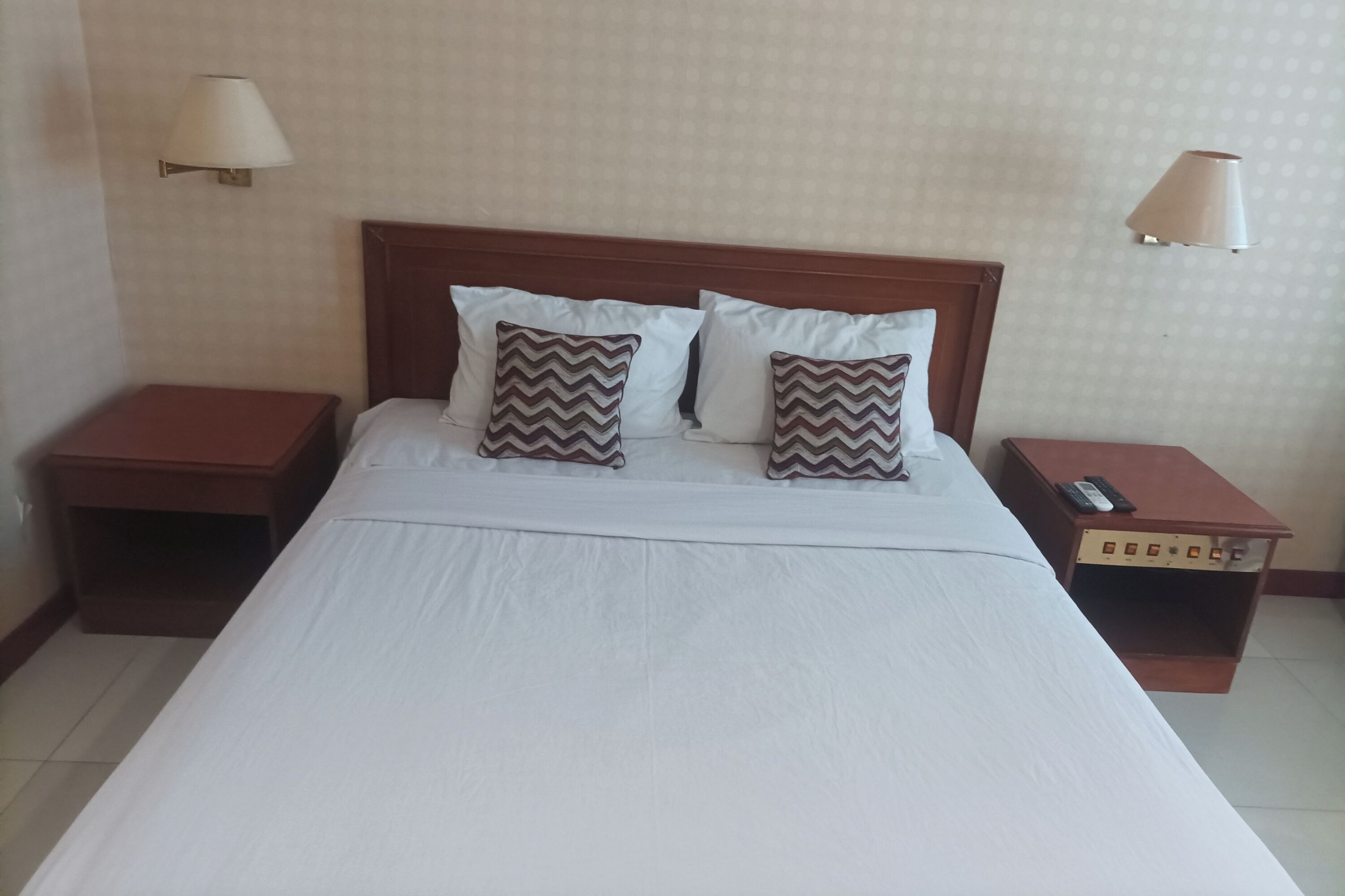 A neatly made bed with two pillows, a wooden headboard, and two bedside tables with lamps and a remote control.