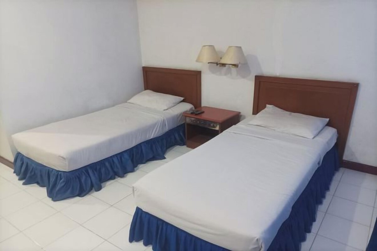 Two single beds with blue skirts, a wooden headboard, a nightstand with a lamp, and a wall-mounted light fixture.