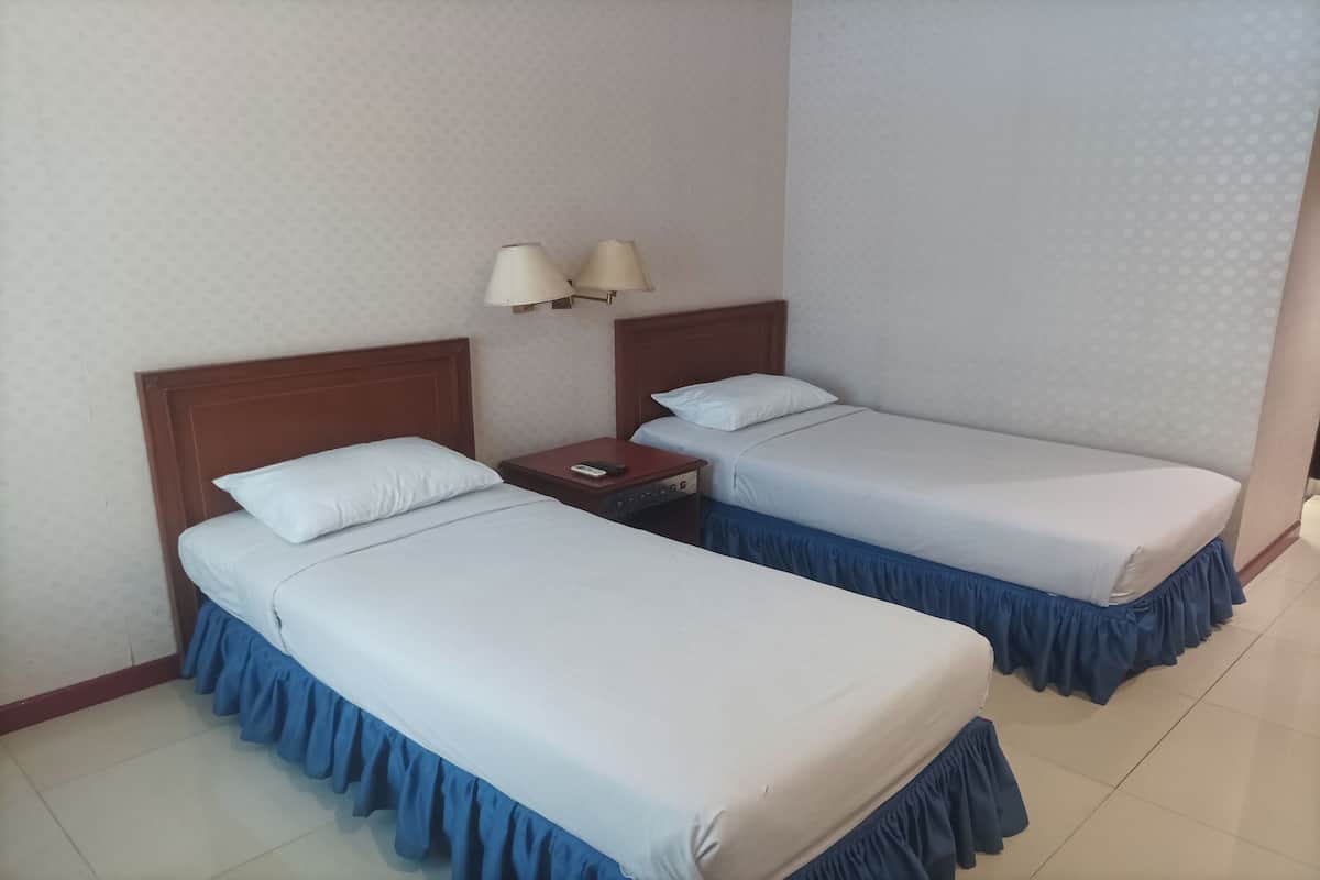 Une chambre d’hôtel avec deux lits simples, chacun doté d’une tête de lit en bois, de draps blancs et d’une housse de couvre-lit bleue. Deux lampes de chevet sont fixées au mur, et une petite table est placée à côté, avec un téléphone et une télécommande.