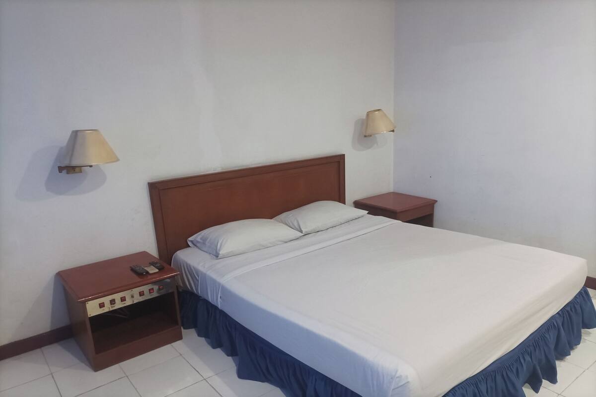 Une chambre à coucher avec une tête de lit en bois, un lit recouvert d’un drap blanc et agrémenté d’une bordure bleue, deux tables de chevet avec des lampes, et une petite table de nuit avec une télécommande.