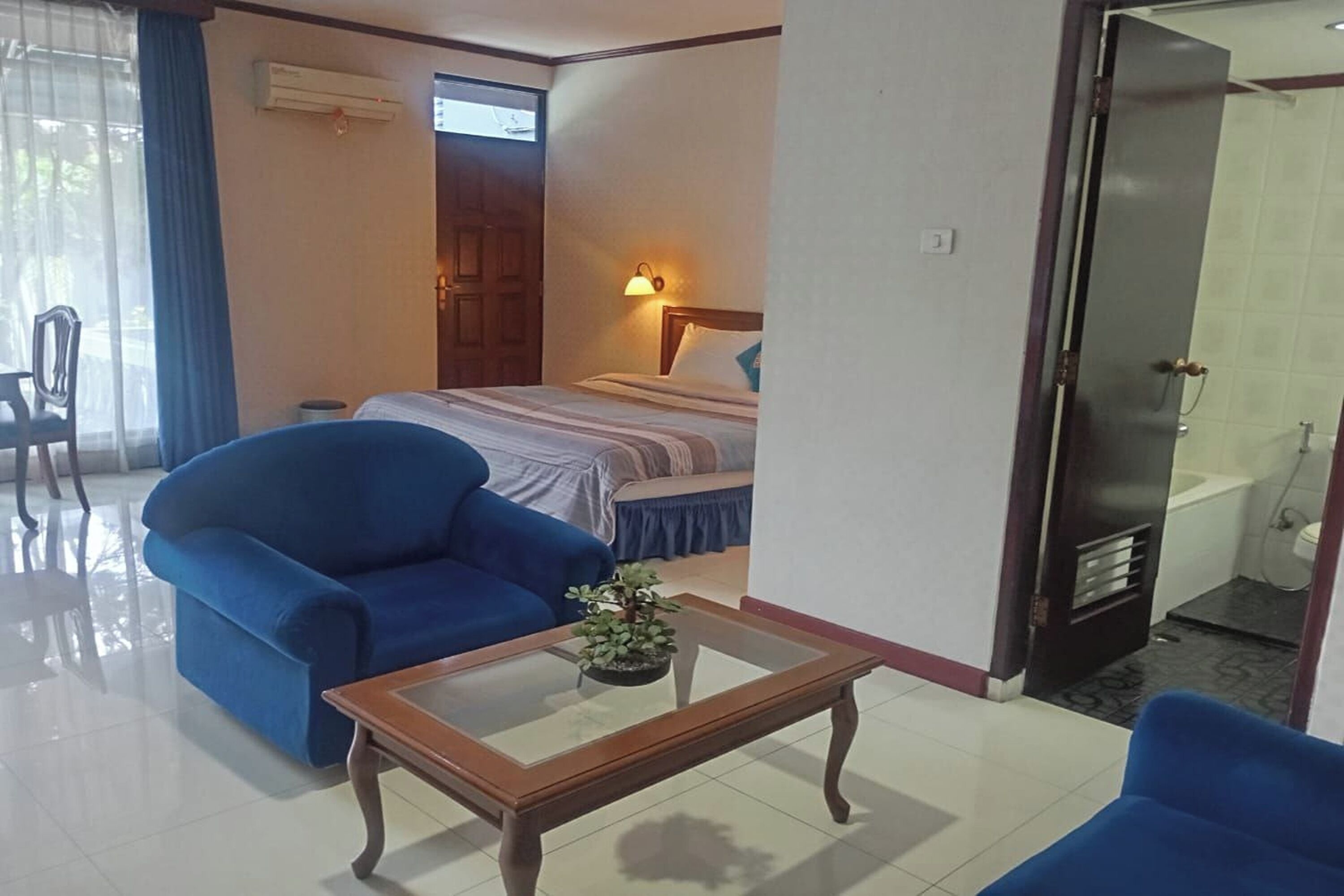Habitación de hotel con cama, sillón azul, mesa de centro de madera con una planta, baño con ducha y puerta que da a otra habitación.