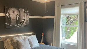 A bedroom with a dark headboard, a bed with white and gray bedding, a guitar, and a window with curtains. - Relaxing 3BR Family Retreat. Spacious • Peaceful • Close to everything (Peralta)