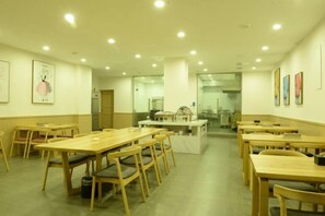 A dining area with multiple wooden tables and chairs, a kitchen in the background, and artwork on the walls.