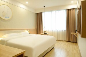 Ein Hotelzimmer mit einem großen Bett, Nachttischen, einem kleinen runden Tisch mit einer Lampe und einem großen Fenster mit Vorhängen.