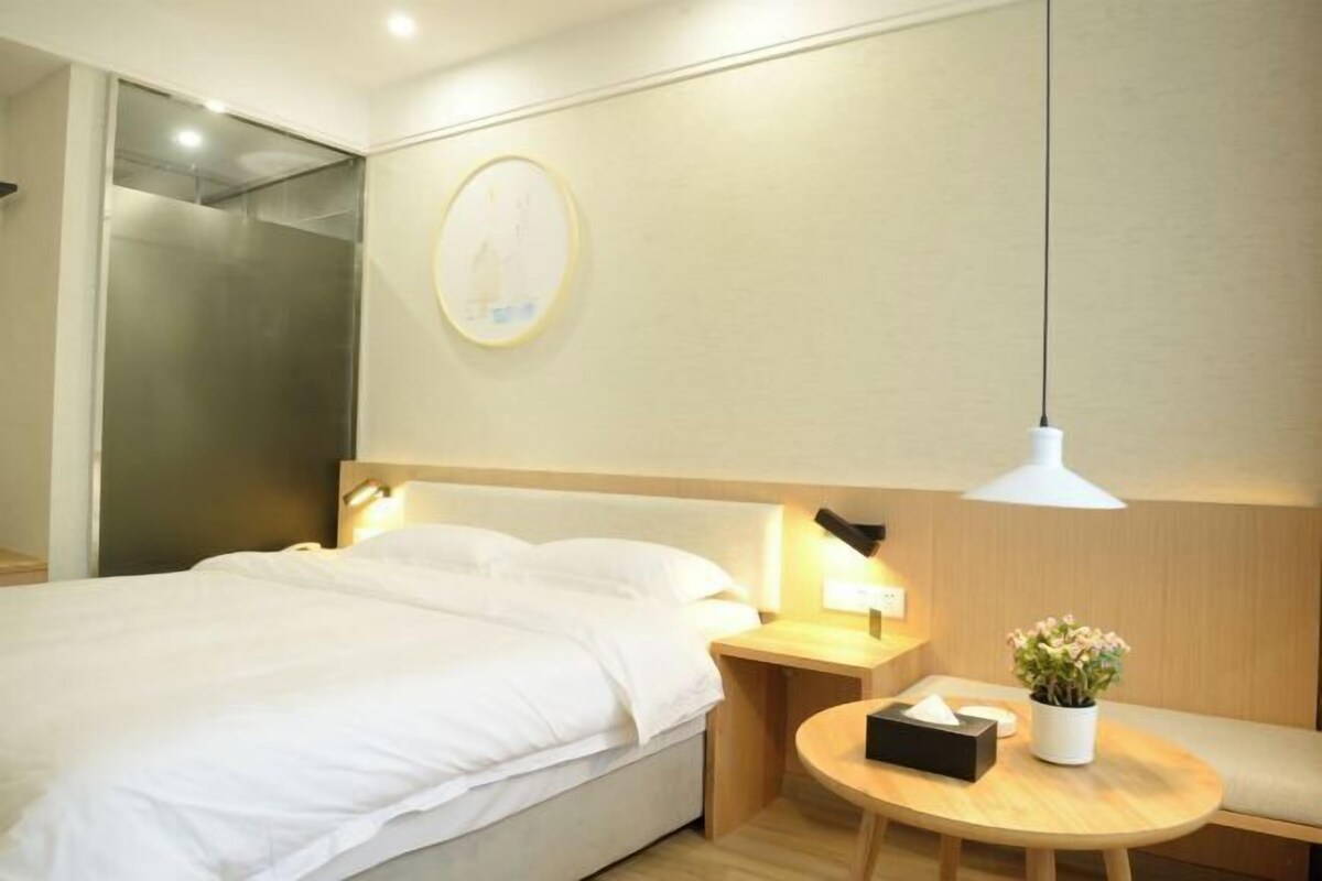 Ein Hotelzimmer mit Bett, Nachttisch mit Lampe, einem kleinen runden Tisch mit Pflanze und einer Wandleuchte.