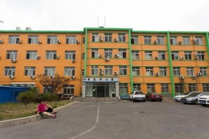 A multi-story building with a green and orange facade, a parking lot, and a person sitting on the ground. - Xiaisi Lexiang Service Apartment (Dalian Xinghai Square) (Dalian)
