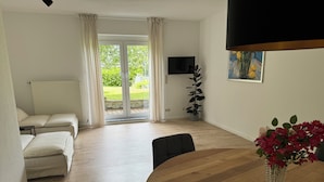 Un salon moderne avec un canapé, une table à manger agrémentée d’un vase de fleurs, un téléviseur fixé au mur et une porte-fenêtre coulissante donnant sur un jardin.