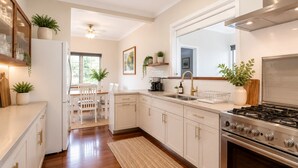 A modern kitchen with white cabinetry, a gas stove, a built-in oven, a dining area with a table and chairs, and a ceiling fan. - Driftwood Cottage (Busselton)