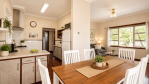 A modern kitchen with white cabinets, a dining area with a wooden table, and a living room with a sofa and large windows. - Driftwood Cottage (Busselton)