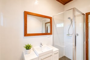 Un baño moderno con un lavabo blanco, un espejo con marco de madera, cabina de ducha y una planta pequeña en el mueble.