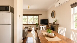 A kitchen with a refrigerator, microwave, and a dining area with a wooden table, chairs, and a television. - Driftwood Cottage (Busselton)