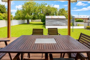 Eine Holzterrasse mit Tisch und Stühlen, ein Rasen, ein Holzschuppen und ein Metallzaun.