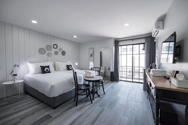 A modern hotel room with a large bed, a small table, a chair, a desk, and a bathroom visible through a glass door.