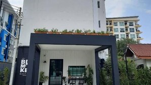 A modern building with a prominent logo on the facade, a covered entrance area with seating, and a clear sky in the background. - Blumonkey Boutique Bangsaen (Chonburi)
