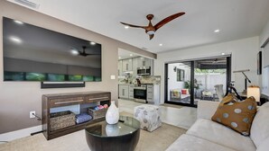 Ein modernes Wohnzimmer mit Flachbildfernseher, Deckenventilator, Glastisch und Sofa mit Punkte-Kissen.