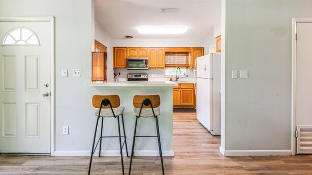 A kitchen with wooden cabinets, a microwave, and a refrigerator. There are two bar stools in front of the kitchen island. - Mid-Century Gainesville Getaway: 3 Mi to UF Campus (Gainesville)