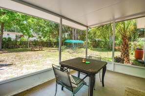 Un patio techado con mesa y sillas, ventanas amplias y vistas a un jardín con césped y árboles.