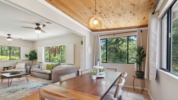 Ein geräumiges Wohnzimmer mit Holztisch, bequemen Sitzmöglichkeiten, großen Fenstern und einem Deckenventilator.