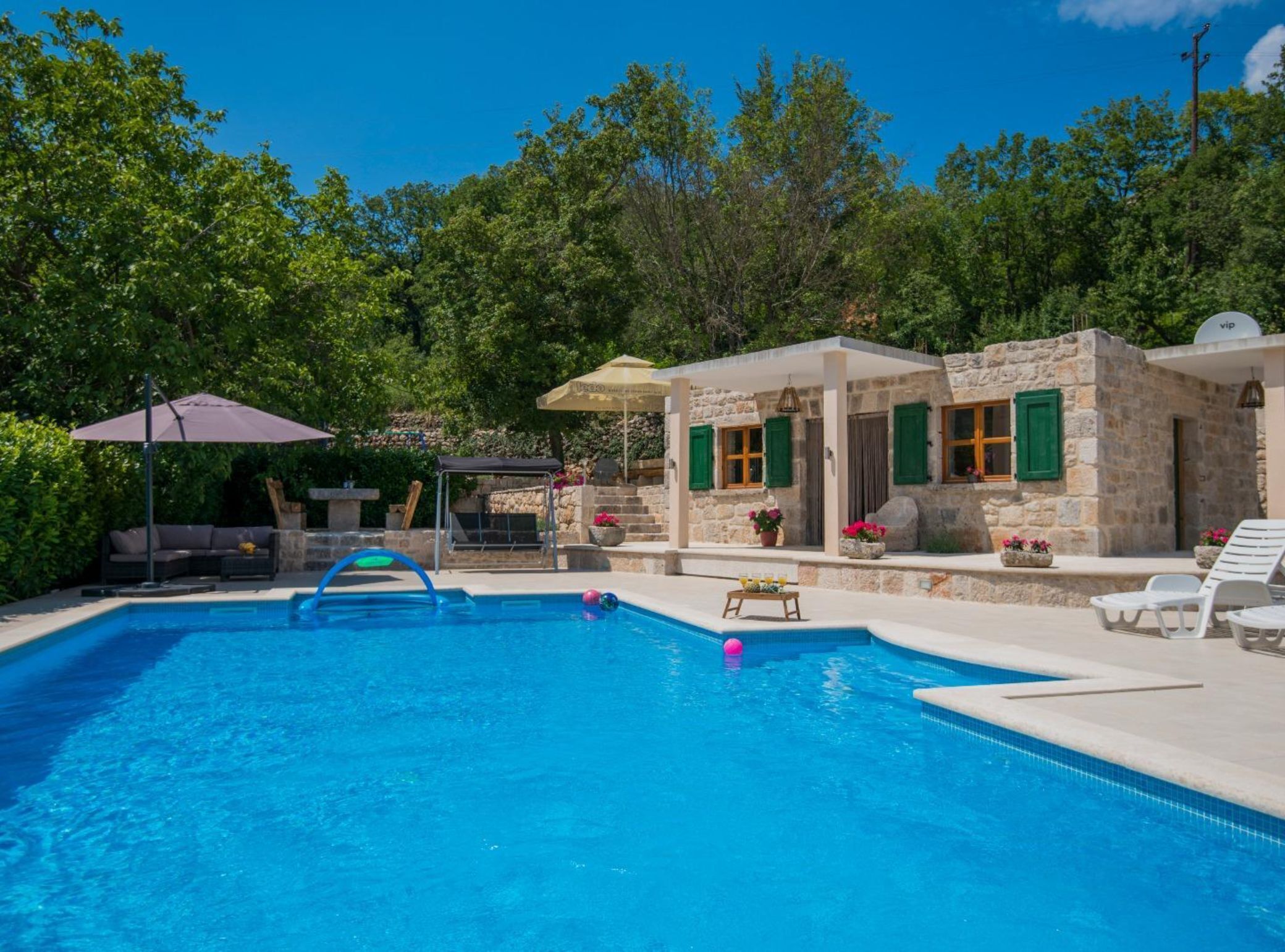 A swimming pool with a stone building, outdoor furniture, and lush greenery.