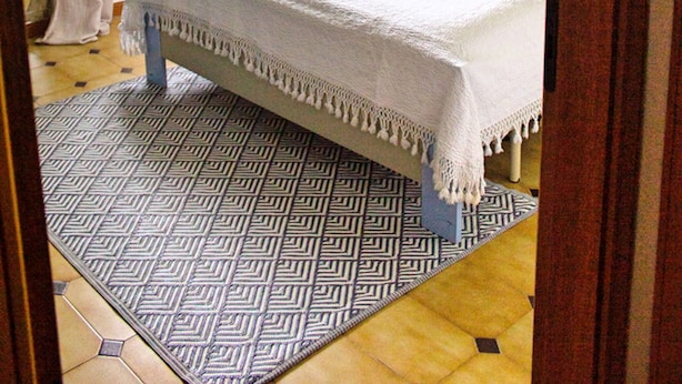 A bedroom with a bed, a window with curtains, a patterned rug, and a wall with a circular decoration. - Blueindaco Sellia Marina - Room Apartment Calabria (Sellia Marina)