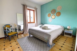 Ein Schlafzimmer mit einem Bett, einem Sessel, einem Nachttisch, einem Spiegel und einem Fenster mit Vorhängen.