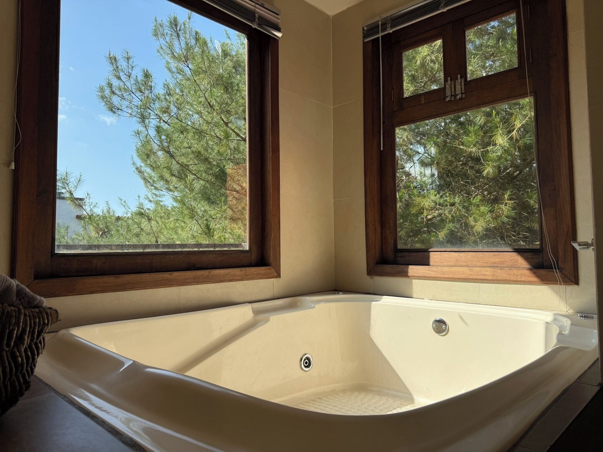 A corner bathroom with a large bathtub, two windows with wooden frames, and a view of trees.
