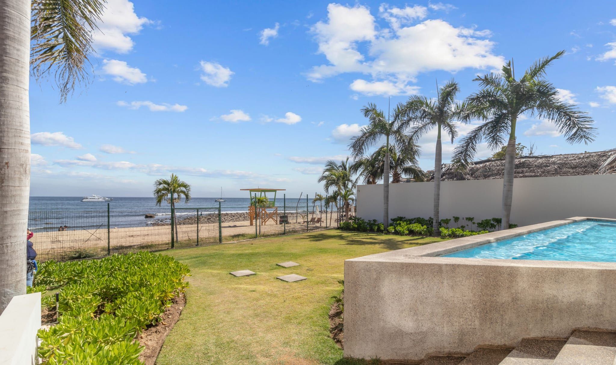 A pool area with a view of the ocean, a fenced garden, and palm trees.