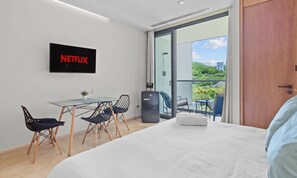 A hotel room with a bed, a small table with chairs, a TV screen displaying 'NETFLIX', and a balcony with a view of greenery and buildings.