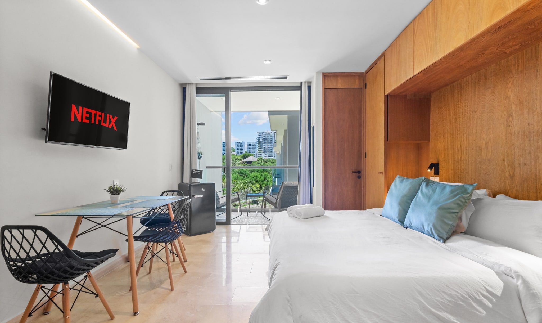 Una habitación de hotel moderna con una cama, un escritorio con una silla, un televisor montado en la pared y un balcón con vistas a la ciudad.