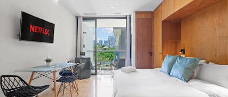 Una habitación de hotel moderna con una cama, un escritorio con una silla, un televisor montado en la pared y un balcón con vistas a la ciudad.