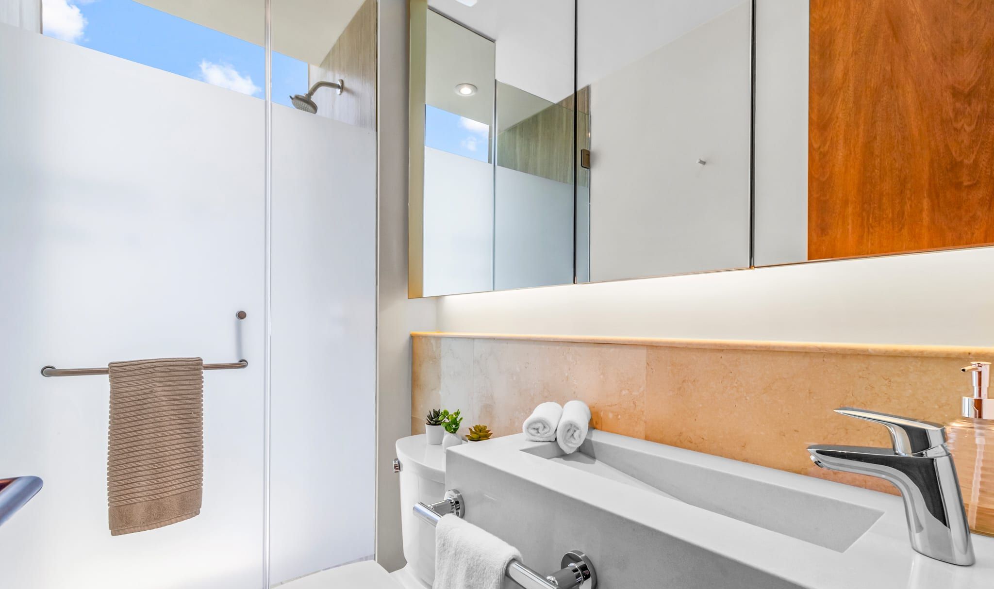 Un baño moderno con mampara de vidrio, un tocador blanco con encimera de mármol y un mueble de madera sobre el espejo.