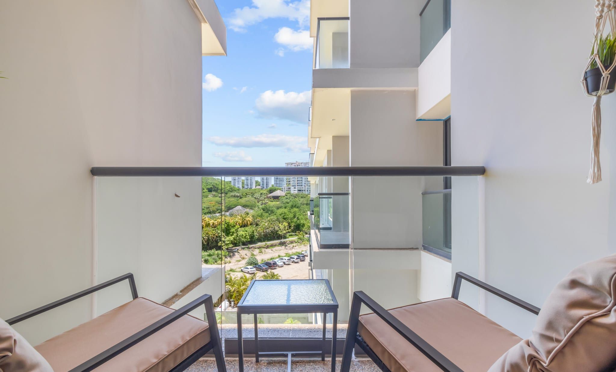 A balcony with a glass table and two cushioned seating areas, offering a view of a landscaped area and buildings in the distance.