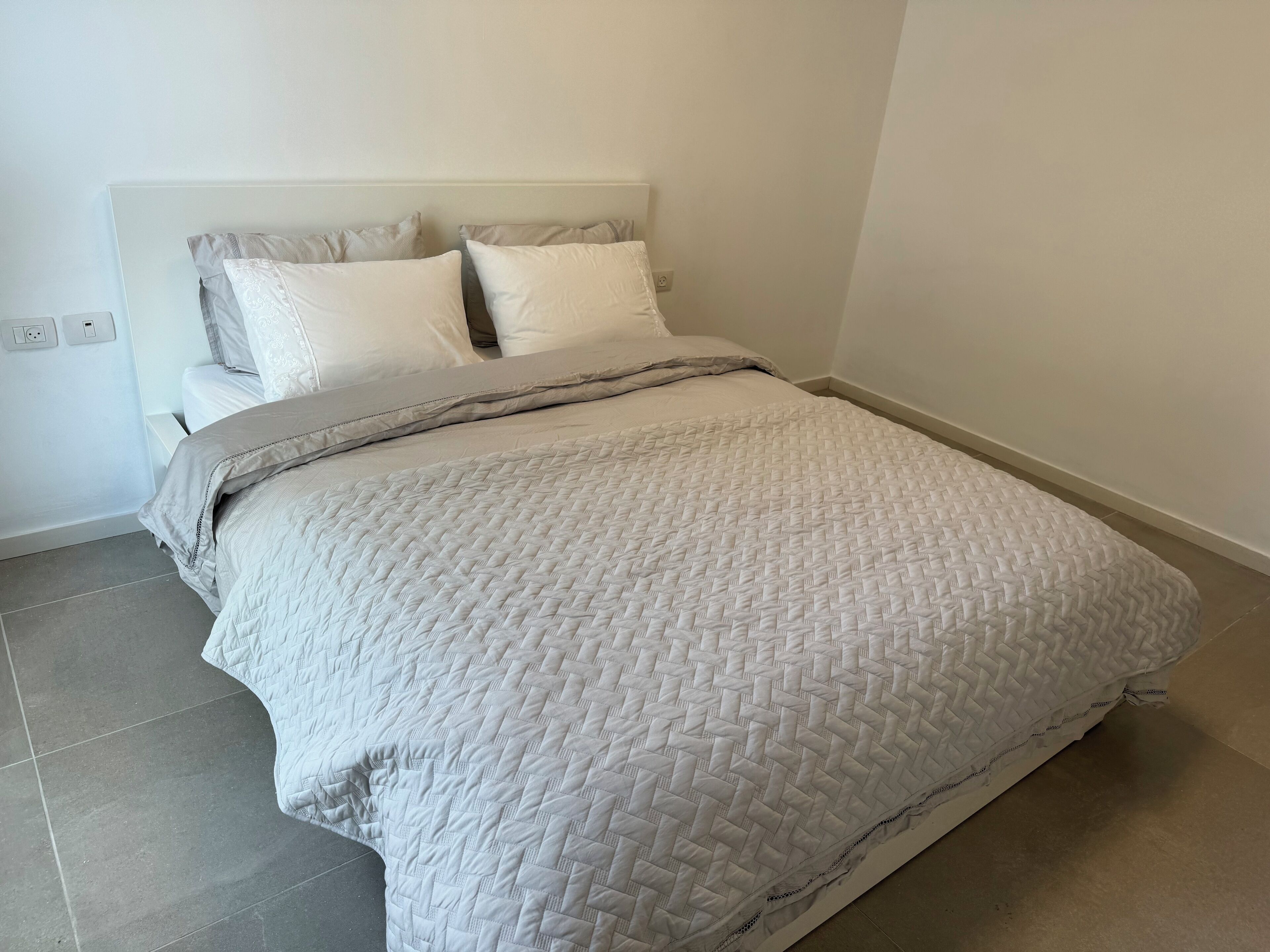 A neatly made bed with a quilted white duvet, a grey blanket, and two pillows. The room has a simple, clean design with a white headboard and a plain wall.
