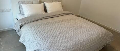 Un lit bien fait, avec une couette blanche matelassée, une couverture grise et deux oreillers. La chambre présente un aménagement simple et épuré, avec une tête de lit blanche et un mur uni.