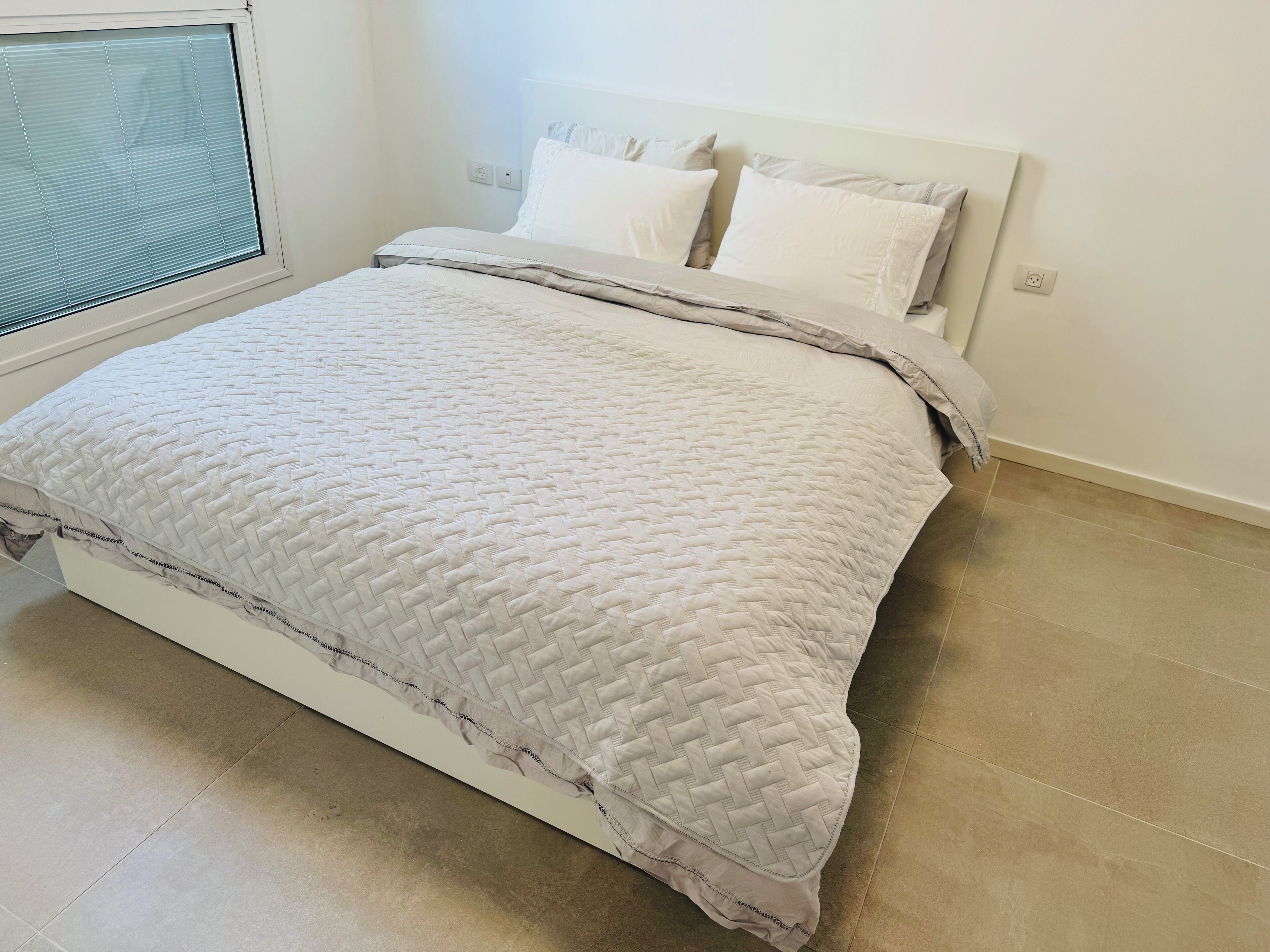 A neatly made bed with a quilted bedspread and pillows, a window with blinds, and a tiled floor.
