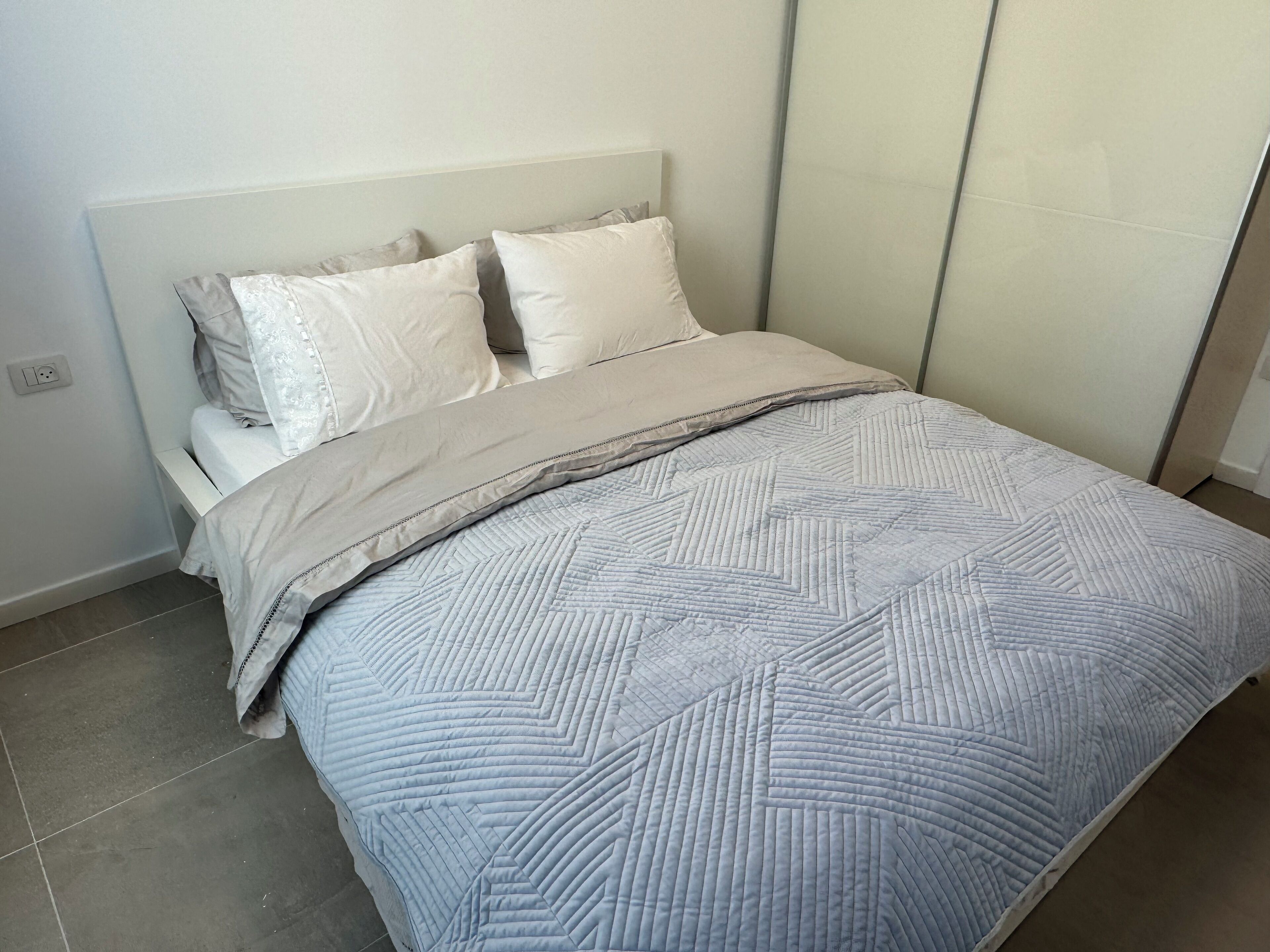 Una cama bien hecha con una colcha estampada y almohadas, una mesita de noche y un enchufe de pared.
