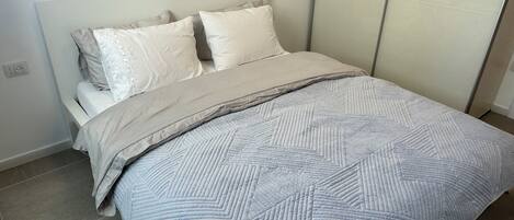 Una cama bien hecha con una colcha estampada y almohadas, una mesita de noche y un enchufe de pared.