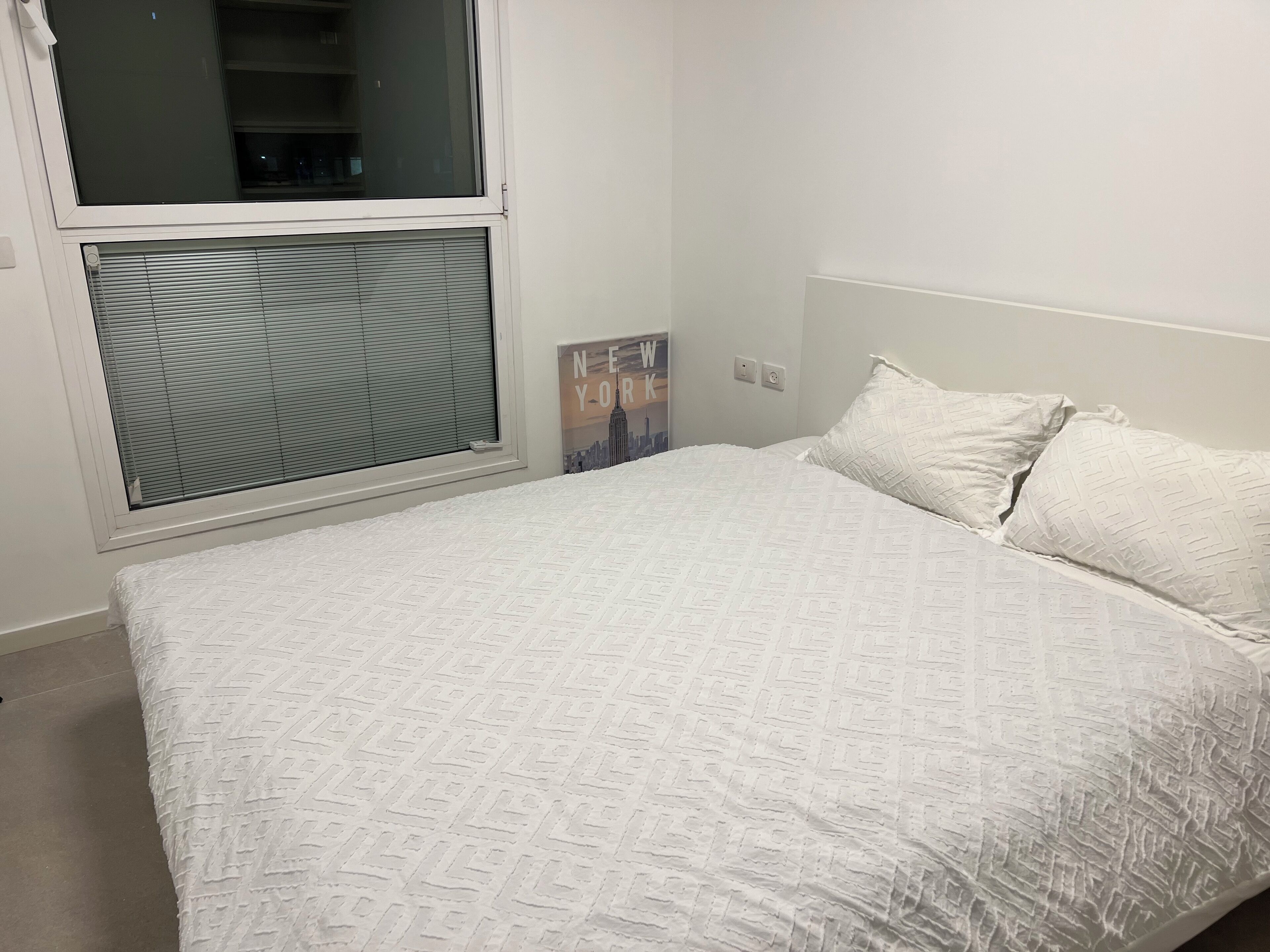 Une chambre à coucher avec un lit, une fenêtre avec des stores, un interrupteur fixé au mur et une affiche accrochée au mur.