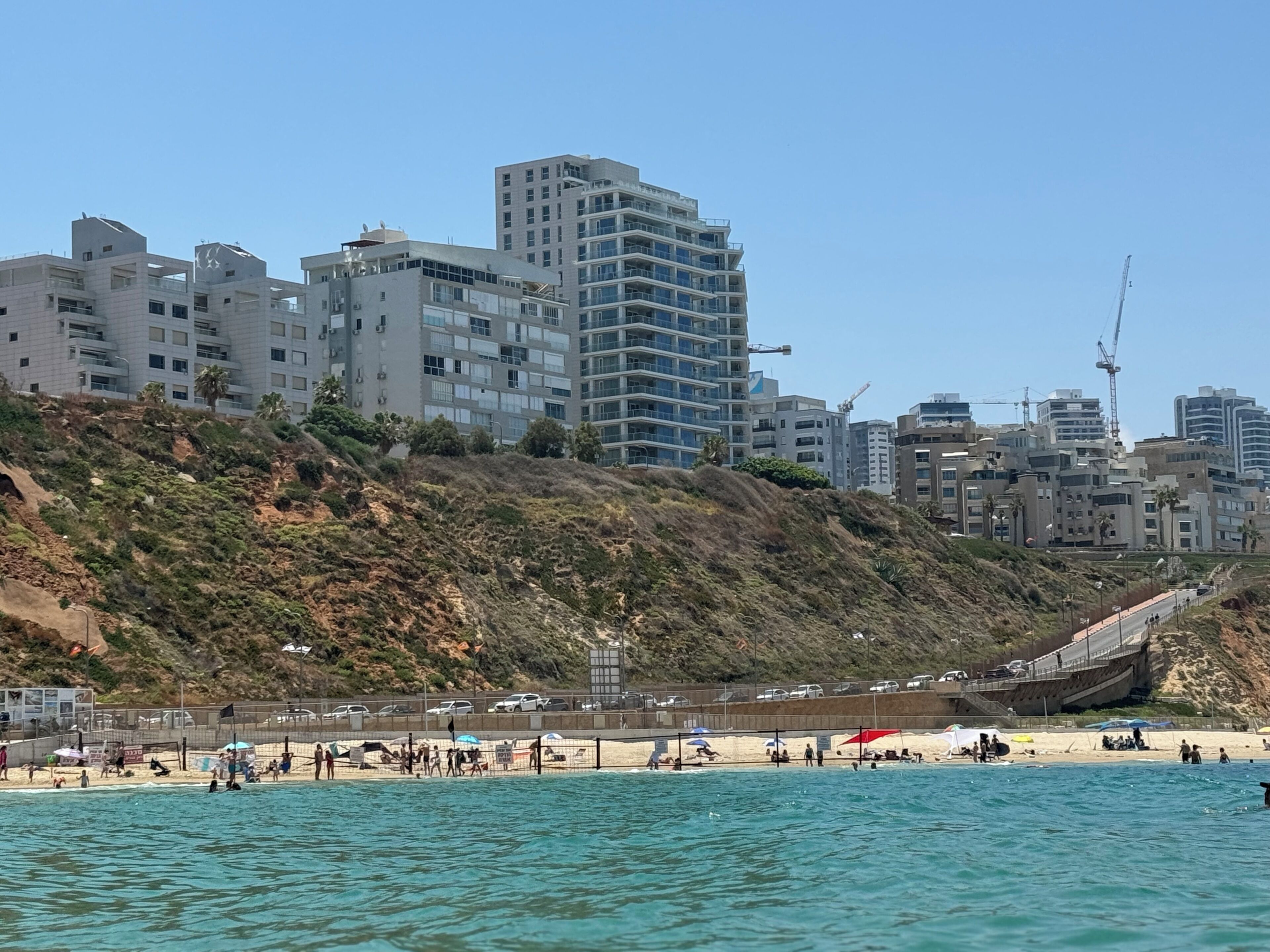 Paisagem costeira urbana com edifícios modernos, uma praia de areia e céu azul límpido.