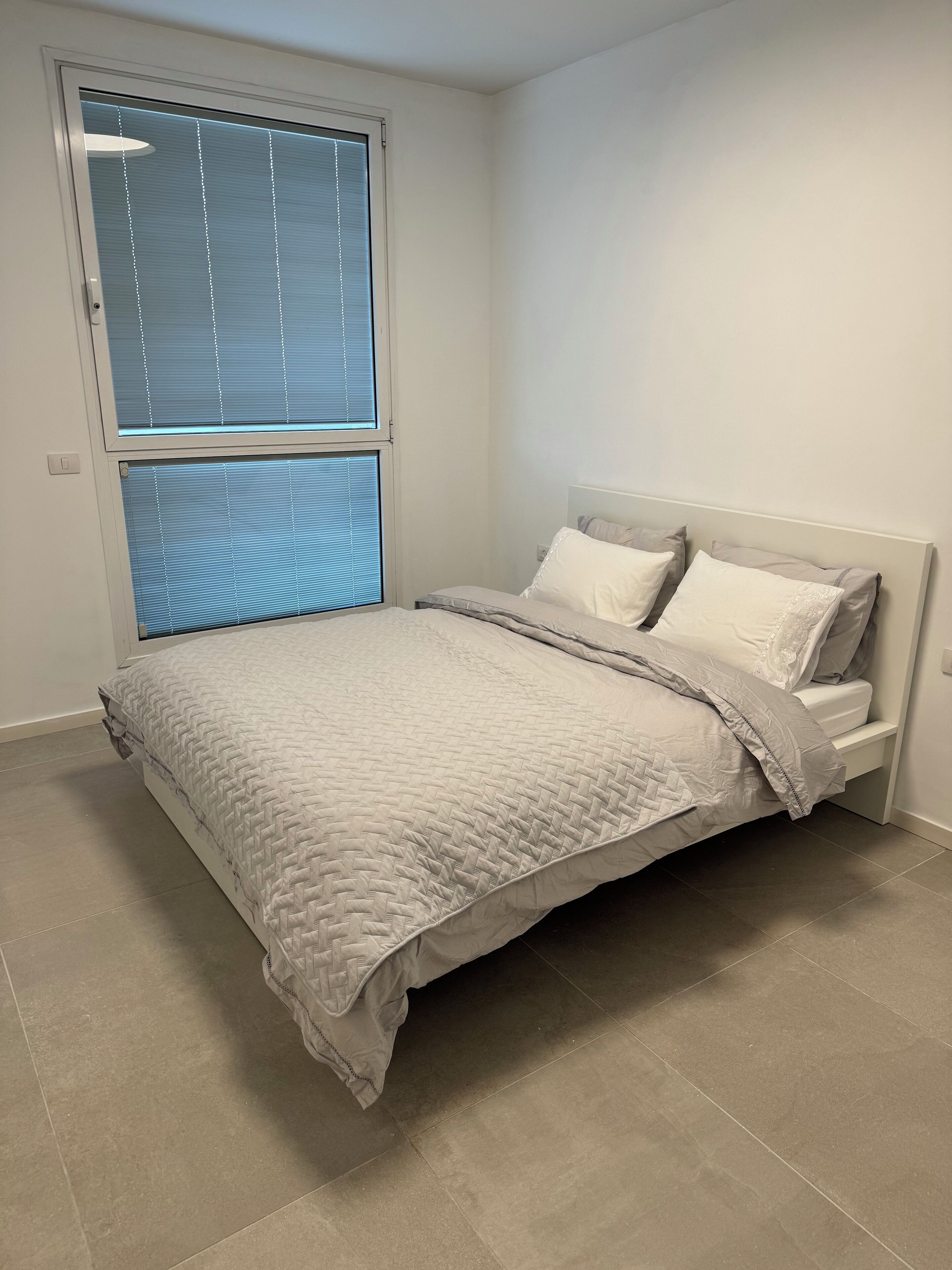 A bedroom with a bed, a window with blinds, and a light switch on the wall.