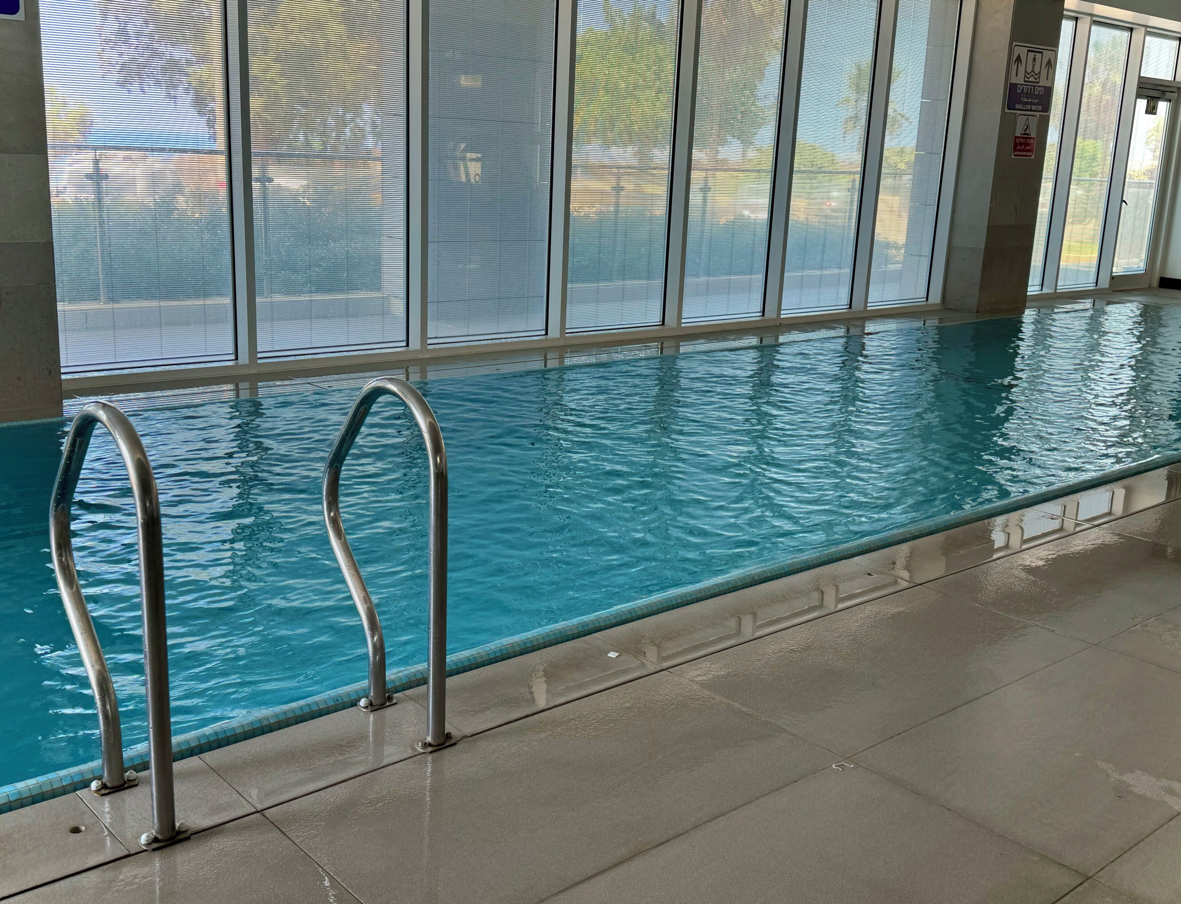 An indoor swimming pool with clear water, metal pool ladders, and large windows.