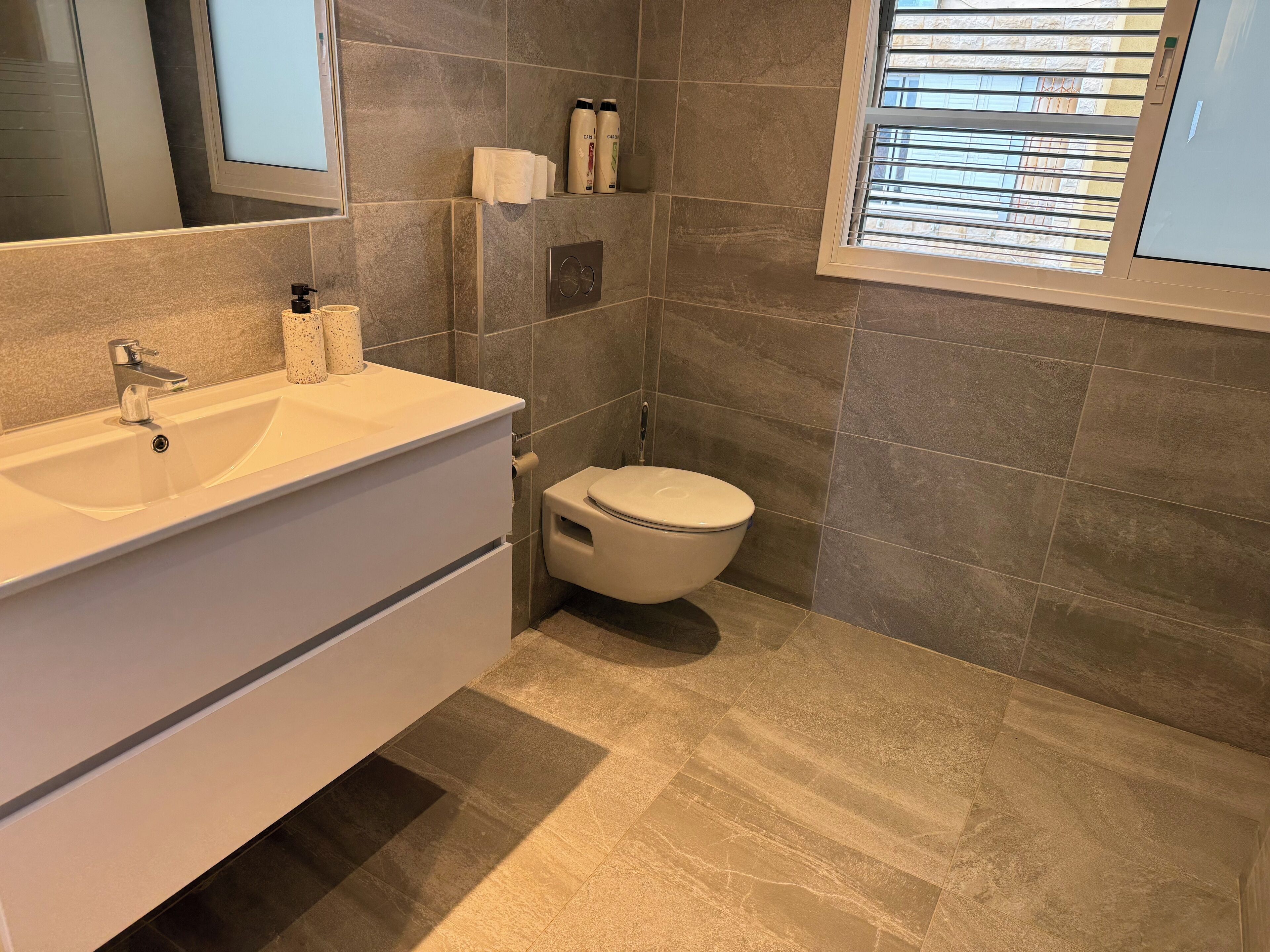 Ein Badezimmer mit weißem Waschtisch, Toilette und einem Fenster mit Jalousien.