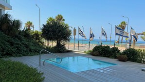 Une terrasse en bois avec une piscine, entourée de verdure et de palmiers, donnant sur une plage où flottent des drapeaux israéliens.